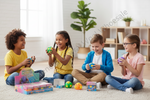 Four children sitting on a white carpet in a bright room, playing with colorful toy building blocks and small balls. A product box labeled 'Raspberry Kettes' is visible on the floor in the foreground.