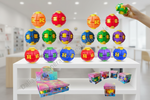 Colorful plastic ball puzzle toys displayed on a white shelf, with a hand holding one toy at the top right. Multiple boxed sets are arranged on the lower shelf, and a blurred indoor room is visible in the background.