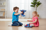 Two children playing with toy trucks in a bright, modern room. A boy in a blue shirt holds a blue and black toy truck, while a girl in a pink dress sits on the floor with a blue and red toy truck.