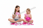 Two young girls sitting on a white studio floor playing with a pink toy makeup set, including a pink handbag, mirror, brushes, and other accessories.