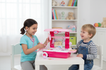 A young girl and a boy are sitting at a small white table playing with a pink toy doctor playset in a bright, white-themed room.