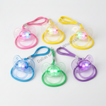 Six pacifiers with attached wrist straps in pink, yellow, blue, green, and purple, each featuring a glowing LED light in the center, arranged on a light gray background.