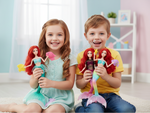 Two children sitting on a white rug in a brightly lit room, each holding a mermaid-themed doll with red hair and a colorful tail.