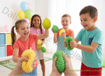 Children playing with green and yellow squishy stress balls in a brightly lit playroom. Multiple kids are shown squeezing the textured balls, with one child in a turquoise shirt on the right and another in a coral shirt on the left.