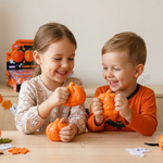 Two children, a girl and a boy, smiling and holding orange plastic jack-o'-lantern toys at a light wood table. The girl wears a beige long-sleeve shirt with a ghost pattern, and the boy wears an orange long-sleeve shirt with a black bat detail.