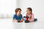A young boy in a blue t-shirt and a girl in a pink hoodie are lying on a white floor, smiling and playing with a small red and black robot toy while holding a remote control in a bright, modern room.