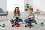 Two children sitting on a carpeted floor in a bright room, each holding a controller next to a small robot toy. The scene includes a gray chair, a window, a plant, and a wooden console table in the background.