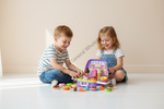 Two children sitting on the floor playing with a colorful toy grocery market set, including a purple market stand and various pretend produce and candy pieces.