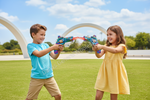 Two children, a boy in a blue shirt and a girl in a yellow dress, playing with colorful toy water guns on a sunny grassy park with white arches in the background.