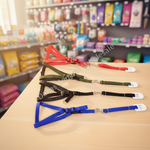 Four pet harnesses in red, green, black, and blue, arranged in a row on a light wood table with a blurred pet store background.