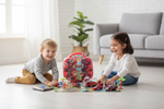 Two children playing with a red toy city set on a light wood floor in a bright living room. A boy in a gray shirt and tan pants is kneeling on the left, and a girl in a white shirt and blue jeans is sitting on the right.