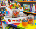 ASMQ Squeeze Soft Stretchy toy shown in its retail display box with a single orange-shaped squeeze toy in front, photographed on a colorful toy store shelf. The packaging features fruit illustrations and the brand name ASMQ.