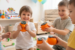 Children smiling and holding bright orange fruit-shaped toy pears in a colorful indoor playroom setting. A large translucent 'Dallas General Wholesales' watermark is visible across the center.