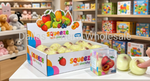 A display of Amino Squeeze Soft Stretchy toy fruit props, including a large box of pale yellow pear-shaped stretchers, a smaller clear box with red and orange fruit designs, and two real-looking pale yellow stretchers being held by a hand on a white table with a toy store background.