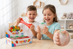 Two children smiling and playing with colorful Squeeze Soft Stretchy toys in a bright room. One child holds a translucent red stretchy ball, while the other holds a clear bubble-filled squeeze toy.
