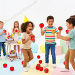 A group of children playing with toy red apples in a brightly lit studio. One child wears a yellow party hat, and another holds a toy apple near the face of another child.