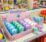A pink retail display box filled with small stretchable axolotl toys in blue, green, purple, and white, shown on a store shelf with other colorful toys in the background.
