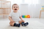 Baby sitting on a carpeted floor wearing socks with a crib and colorful toys in the background.