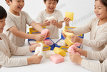 Children sitting around a table squeezing colorful squishy soap bars from open blue and yellow boxes. The soaps are pink, white, and yellow, and a translucent watermark reading ' wholesale' is visible across the scene.