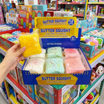 A hand holds a yellow Butter Squishy toy in front of an open retail display box of 12 colorful Butter Squishies in a toy store aisle.