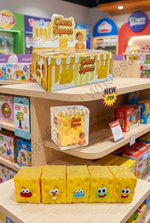 A retail display of yellow sponge-shaped squishy toys labeled 'Cheese Squeezies' on wooden shelves, with a larger box on top and five individual squishys lined up in front. The toys have different colored faces, and the background shows a colorful toy store interior.