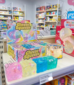 A retail display box and several translucent squishy cubes labeled 'Cheese Squeeze' on a store shelf, with a hand holding one of the squishies in the background.