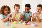 Four children sitting at a table smiling and playing with colorful squishy stress balls and slime on a light wood surface against a plain off-white wall.