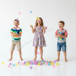 Three children laughing and playing with pastel ball bearings in a bright white studio setting. The child on the left wears a striped shirt and khaki shorts, the center child wears a floral dress, and the child on the right wears a gray and pink graphic T-shirt with blue denim shorts.