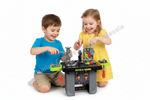 Two children, a boy in a blue shirt and a girl in a yellow polka-dot dress, playing with a toy mechanic workbench on a white background. The workbench has toy tools and parts, and there is a diagonal 'Dallas Goods Wholesale' watermark across the image.