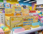 Must Baby Crispy Butter Stick Squishy in coconut oil, shown in a yellow retail display box, a single stick package, a stack of three colored butter sticks, and a hand holding a blue-tinted stick.