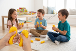Three children sitting on a rug in a bright living room, each holding a yellow corn-shaped toy. A pair of hands in the foreground is squeezing a matching yellow corn toy.