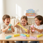 Three children sitting at a table in a bright room, each holding a colorful rectangular toy block labeled "BUTTER." One child stretches a yellow block, while others are held in pastel blue, pink, and yellow.
