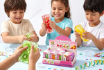 Three children are happily squeezing colorful ice squishy toys shaped like strawberries, kiwi, and yellow fruit, gathered around a pink display box labeled 'ice Squishy' on a light blue table with confetti and ice cubes.