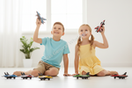 A young boy in a light blue t-shirt and a girl in a yellow dress sit on the floor playing with toy fighter jets. Several colorful toy jets are lined up on the floor in front of them, with a bright, sunlit room in the background.