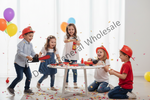Five children playing with toy firefighter-themed items around a small table, including red helmets, a toy chainsaw, a fire extinguisher, and colorful balloons in a bright indoor setting with confetti.