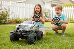 Two children playing with a large white and black remote-controlled monster truck on a green lawn, with a white picket fence and house in the background. The children are holding steering wheels.