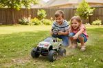 A young boy and girl crouching on a green grass backyard while playing with a camouflage-patterned remote control monster truck.