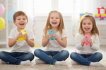 Three children sitting on the floor holding colorful toys in a bright room.