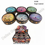 Six round Halloween-themed glitter toy containers with eyes and smiling faces, shown above a retail display box. The tops are decorated in different colors: blue, pink, green, orange, clear, and purple, with holographic glitter visible in some.