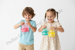 Two children, a boy in a light blue t-shirt and a girl in a cream floral dress, smiling and playing with translucent plastic bags filled with pink, blue, and yellow glitter, set against a plain white background.
