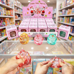 A retail display and hands demonstrating squishy jelly cake toys. In the foreground, two hands squeeze pink and red squishy cake toys, while in the background, a pink 'Happy Cake Squishy' branded display stand holds multiple boxed and loose versions of the toy in pastel colors.
