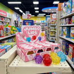 A retail display box of Jumbo Lips jelly lip candies on a store shelf, with several colorful jelly lips arranged in front. The display is pink and white with the words 'JUMBO LIPS' on the sign, and the surrounding shelves are filled with toys and other products in a toy store.