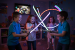 Three children playing with glowing LED toy swords in a colorful children's room. The two boys in blue shirts are holding the swords that glow green and pink, while a girl in a white patterned shirt stands in the background.