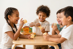 Three children sitting around a small wooden table, laughing and playing with a toy cheese game. A box labeled Texas Toy Company is visible on the table, with a Dallas General Wholesale watermark across the center.