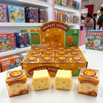 Squeeze Cheese toy set displayed on a store shelf, including a large display box, two small packaged cubes, and two loose cheese-shaped pieces in the foreground. The toys are yellow with printed holes, and the background shows colorful toy store shelves with other products.