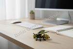 A small yellow and black toy bee figurine sits on a light wood desk in front of a blurred computer monitor, keyboard, mouse, and open notebook. A diagonal 'Dallas General Wholesale' watermark overlays the scene.