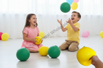 A young girl in a pink dress and a boy in a yellow shirt sit on a white floor, laughing and reaching for colorful balloons in a bright room. A hand in the foreground holds a yellow balloon.