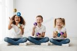Three children sitting cross-legged on a white carpet in a bright room, laughing and juggling shiny colorful balls. They are wearing white t-shirts and blue jeans.