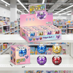 A retail display box and individual packaging for 'Decompress Rotate Ball' fidget balls are shown on store shelves, with four colorful balls in blue, purple, gold, and red displayed in front. The background is a blurred toy store aisle.