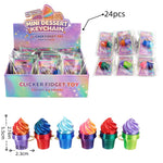 A bulk display box of mini dessert keychain clicker fidget toys, shown alongside individual packaged units, a total count of 24 pieces, and a row of six colorful toy examples. The packaging features a holographic purple and pink design, and the toys have colorful swirl tops resembling ice cream or frosting.
